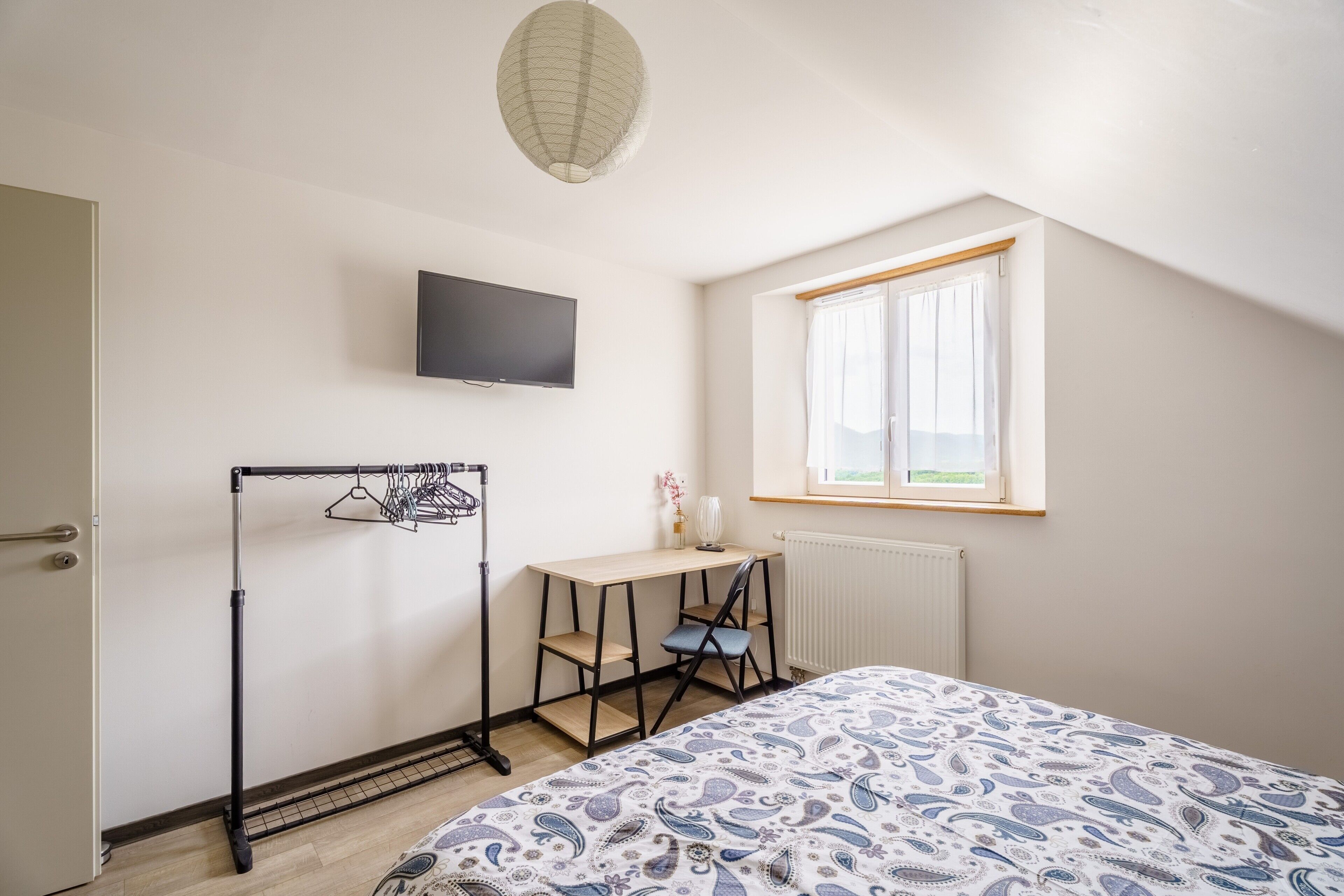 Een slaapkamer met een bed, bureau, stoel, televisie, raam en een radiator.