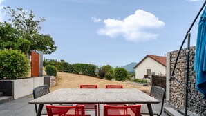 Een terras buiten met een tafel en stoelen, omringd door groen en uitzicht op een berg.