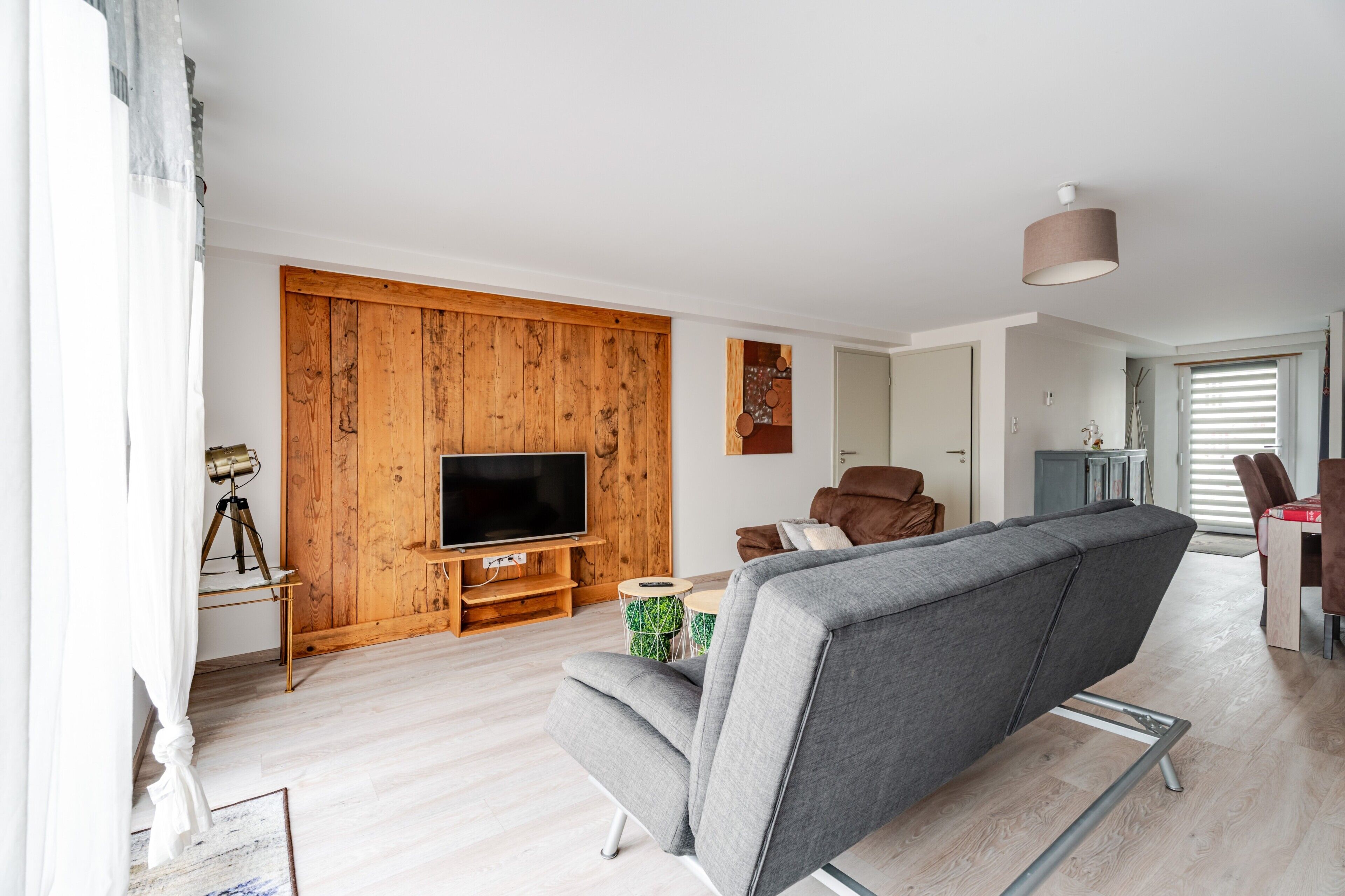 A modern living room with a wooden accent wall, a flat-screen TV, a grey sofa, and a dining area with a table and chairs.
