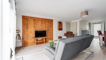 A modern living room with a wooden accent wall, a flat-screen TV, a grey sofa, and a dining area with a table and chairs.