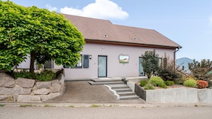 Een huis met twee verdiepingen, een stenen muur, een groene struik en een berg op de achtergrond.