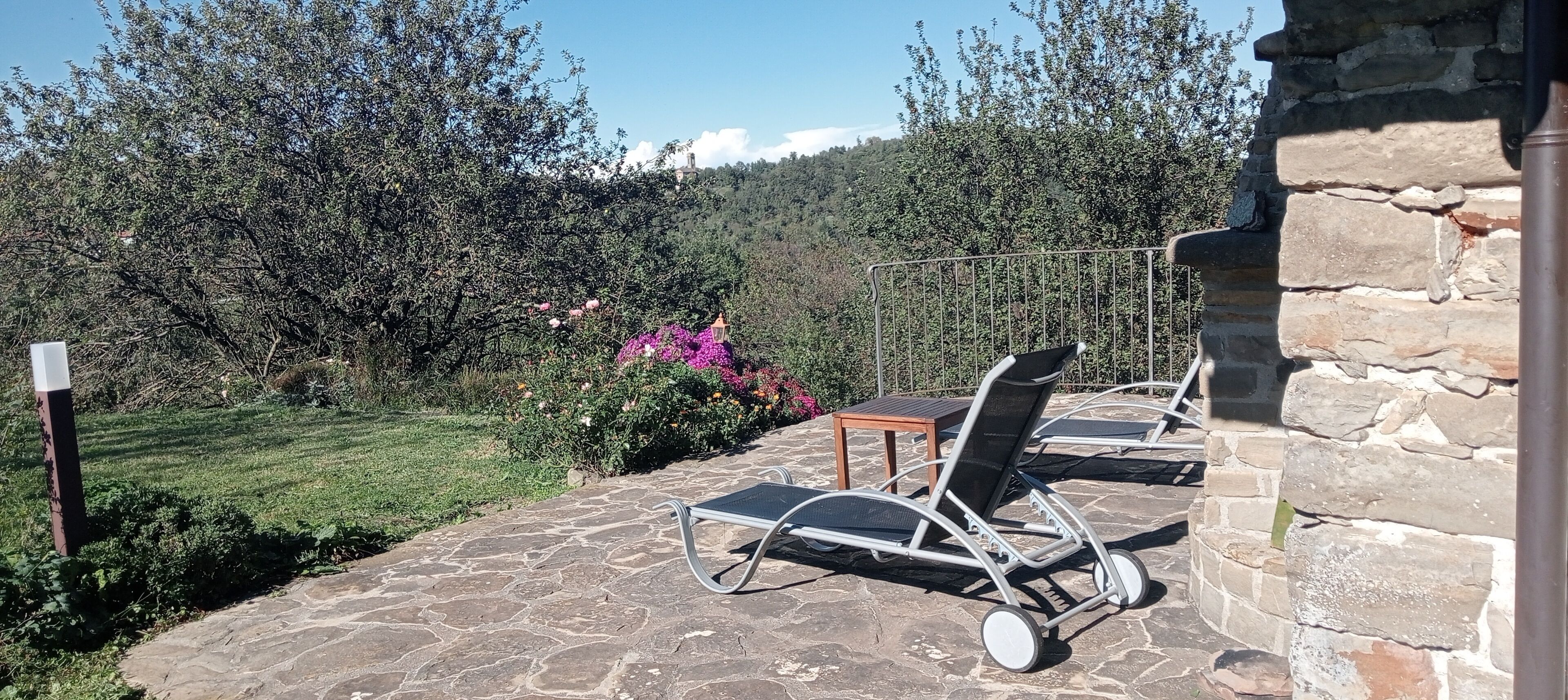 Eine Steinterrasse mit Außenmöbeln, eine Steinmauer und ein Garten mit Sträuchern und Blumen.
