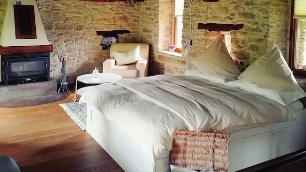 A bedroom with a stone wall, wooden beams, a bed, a chair, a small table, and a fireplace. - Charming Historic House in Provincia di Cuneo, Italia (Mombarcaro)