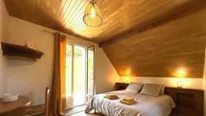 Un dormitorio con techo de madera, una cama con almohadas y una manta, una mesita de noche y una ventana con cortinas.