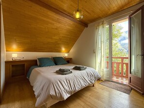 Une chambre avec un plafond en pente, un lit recouvert dâune courtepointe blanche, deux oreillers, une table de chevet avec une lampe, et une vue sur la verdure Ă travers une porte-fenĂȘtre coulissante.