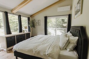 A bedroom with a bed, a dresser, a window with curtains, a plant, and an air conditioning unit.