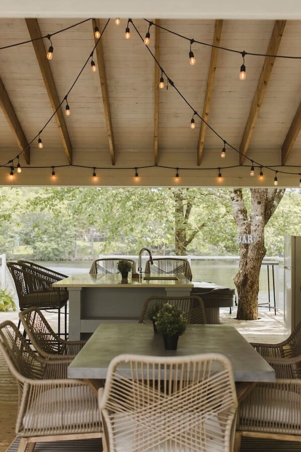 An outdoor dining area with a wooden ceiling, string lights, wicker chairs, and a view of a lake.