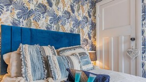 A bedroom with a blue headboard, multiple pillows, a patterned wall, a door with a heart-shaped decoration, and a bedside lamp. - 4 Bed in Whitby (oc-w42084) (Whitby)