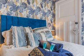 Un dormitori amb un capçal blau, molts coixins, un mur amb un disseny i una porta decorada amb un cor, i una làmpada de nit.