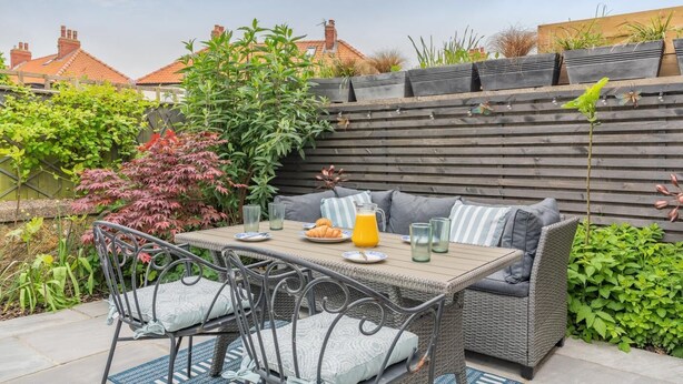A patio with a table set for two, surrounded by wicker furniture and plants. - 4 Bed in Whitby (oc-w42084) (Whitby)