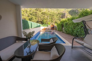 A pool area with wicker furniture, a glass table, and a view of a mountainous landscape.