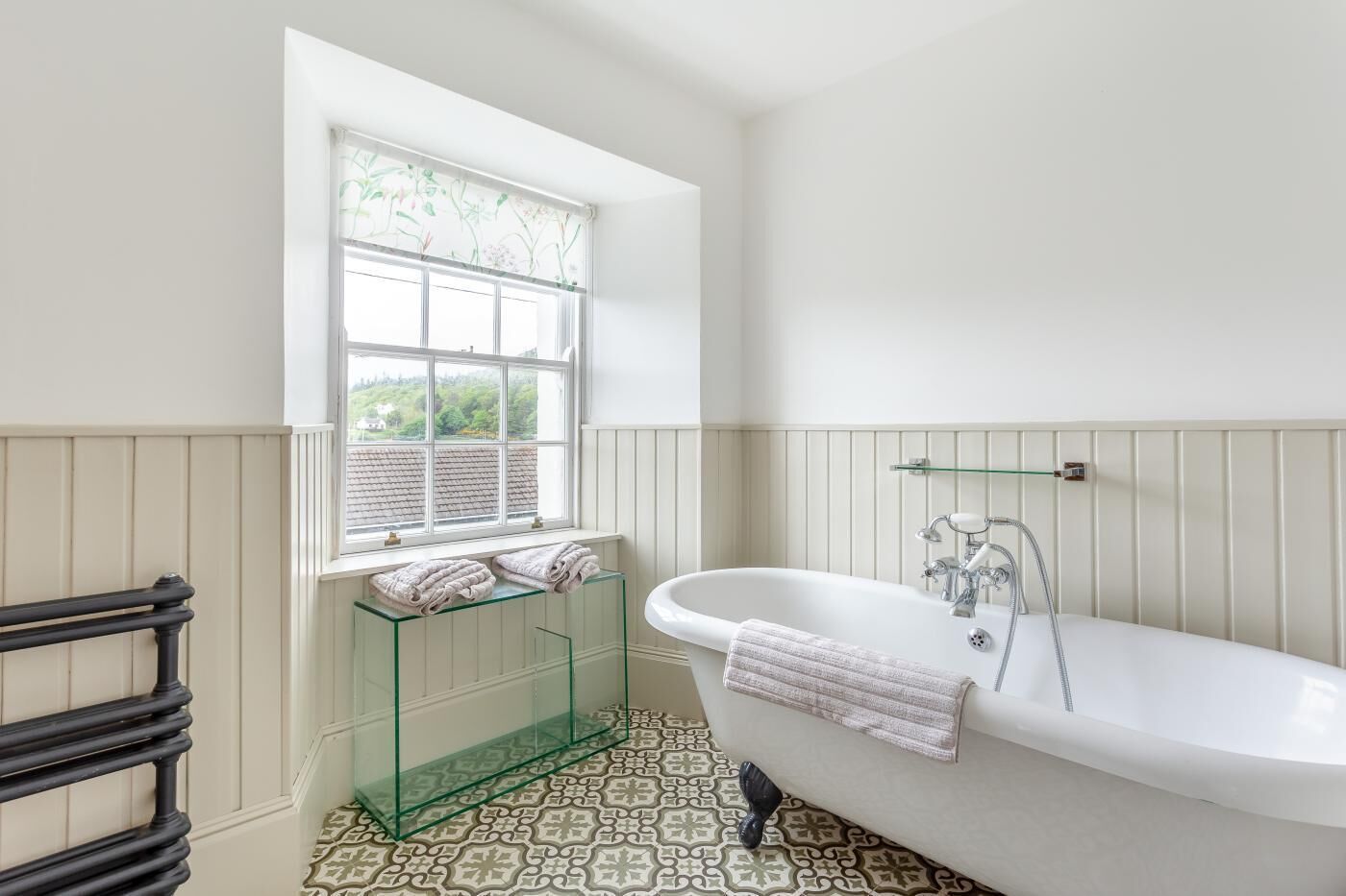 A bathroom with a white bathtub, a glass shelf, a window with a view of greenery, and patterned floor tiles.