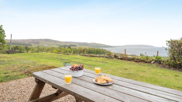 A picnic table with a bowl of grapes, a glass of orange juice, and a plate of croissants, overlooking a coastal landscape. - 5 Bed in Machrie (oc-h42798) (Machrie)