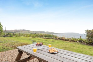 A picnic table with a bowl of grapes, a glass of orange juice, and a plate of croissants, overlooking a coastal landscape. - 5 Bed in Machrie (oc-h42798) (Machrie)