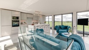 Een modern interieur met een glazen eettafel, een bank, een keuken met witte kastjes en een groot raam met uitzicht op het landschap.