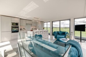 A modern interior with a glass dining table, a sofa, a kitchen with white cabinets, and a large window with a view of the countryside. - 1 Bed in Claxby St. Andrew (oc-r42838) (Claxby St. Andrew)