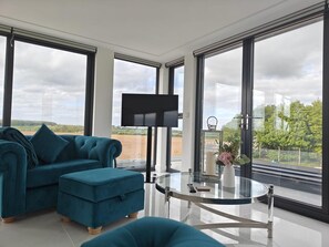 Una sala de estar moderna con un sofá color turquesa, un reposapiés y una mesa de centro de vidrio. Un televisor de pantalla plana está montado en la pared, y amplias ventanas ofrecen vistas al campo.