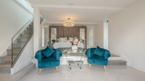 Een moderne woonkamer met een glazen salontafel, twee teal fauteuils, een kroonluchter en een trap naar een slaapkamer.