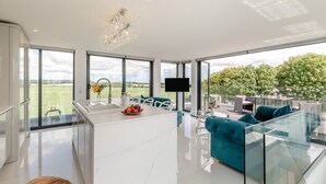 Een moderne keuken met een groot eiland, een zithoek met blauwgroene banken en uitzicht op een groen landschap door grote ramen.