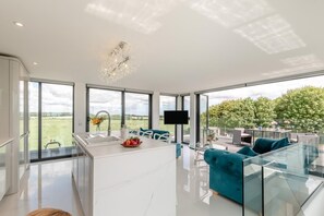 A modern kitchen with a large island, a dining area with teal sofas, and a view of a green landscape through large windows. - 1 Bed in Claxby St. Andrew (oc-r42838) (Claxby St. Andrew)