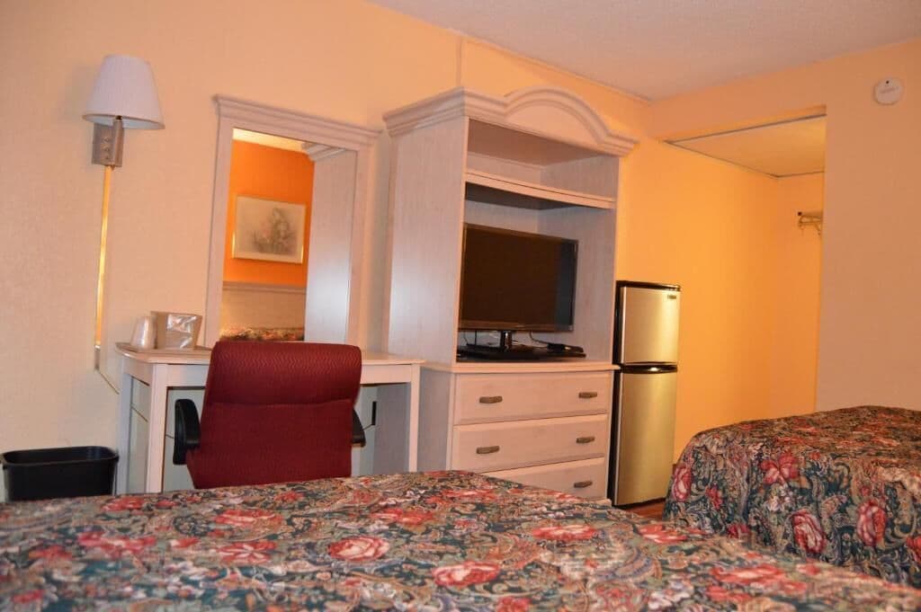 Une chambre d’hôtel équipée d’un bureau, d’une chaise, d’une télévision, d’un réfrigérateur et de deux lits recouverts de couvre-lits à motifs floraux.