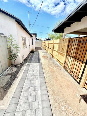 Un estrecho sendero pavimentado entre dos edificios, una valla de madera y un pequeño árbol.