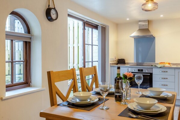 Un comedor con una mesa preparada para cuatro, una cocina al fondo y un reloj en la pared.