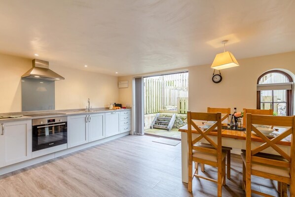 A modern kitchen with a built-in oven, a dining area with wooden chairs, and a view of a wooden deck. - 3 Bed in Lanreath (oc-j42768) (Lanreath)