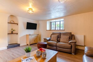 A living room with a brown leather sofa, a wooden coffee table, a television mounted on the wall, and a window with a view of greenery. - 3 Bed in Lanreath (oc-j42768) (Lanreath)