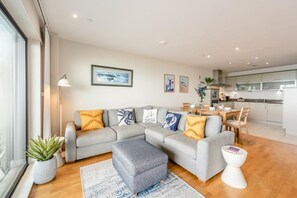 Ein modernes Wohnzimmer mit einem grauen Ecksofa, einem kleinen Hocker, einem Esstisch mit Stühlen, einer Küchenzeile und einem großen Fenster.