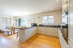 A modern kitchen with a central island, wooden flooring, and a dining area with a bench. - 3 Bed in Widemouth Bay (oc-g42836) (Widemouth Bay)