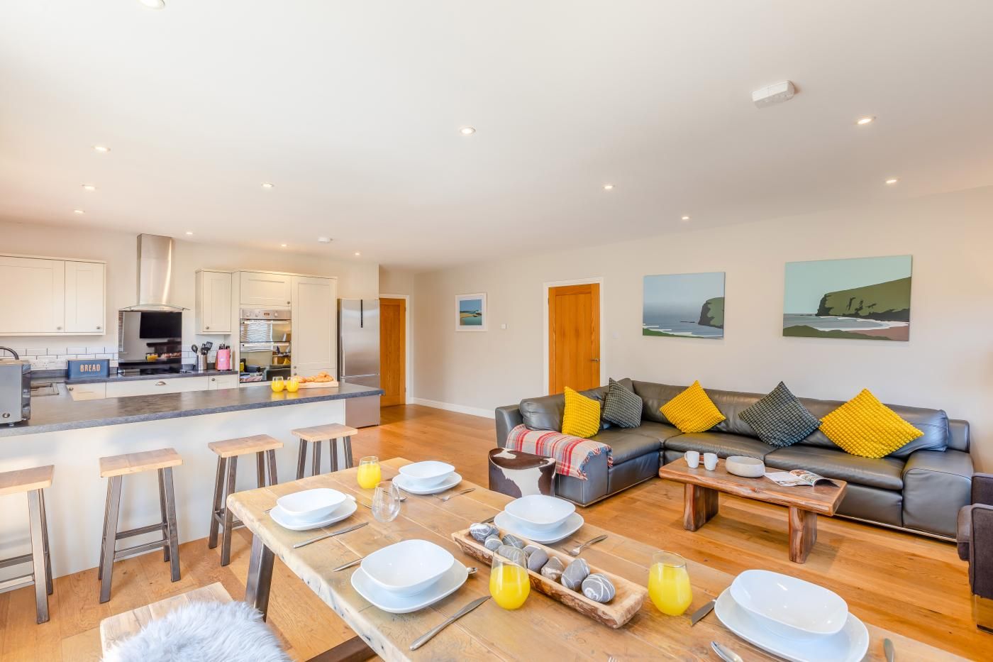 A modern kitchen with a central island, a dining area with a wooden table, and a living room with a sofa and cushions.