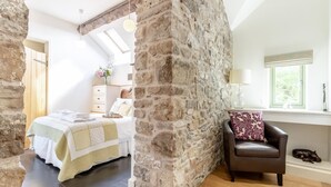 A bedroom with a stone wall, a bed with a quilt, a dresser, a window with a view, and a leather chair with a cushion. - 6 Bed in Llansantffraid (oc-r42827) (Llansantffraid)