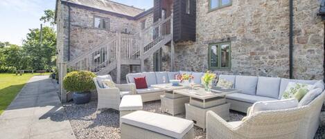 A stone building with a staircase, outdoor seating area with wicker furniture, and a garden path.
