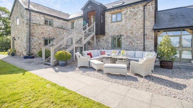 A stone building with a staircase leading to a second floor, a covered outdoor seating area with a sofa and coffee table, and a small tree in a pot. - 6 Bed in Llansantffraid (oc-r42827) (Llansantffraid)
