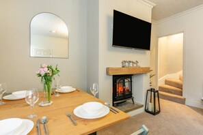 Un comedor con una mesa de madera preparada para cuatro personas, un florero con flores, una chimenea y un televisor montado en la pared.