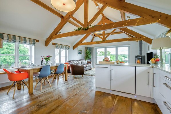 A spacious kitchen with wooden beams, a dining area with colorful chairs, and a large window overlooking greenery. - 3 Bed in Marhamchurch (oc-h29904) (Marhamchurch)