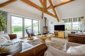 A cozy living room with a fireplace, comfortable seating, a television, and large windows. - 3 Bed in Marhamchurch (oc-h29904) (Marhamchurch)