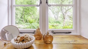 Una habitación con un ventanal grande, una mesa de madera, un cuenco blanco con plantas secas y dos jarrones de vidrio.