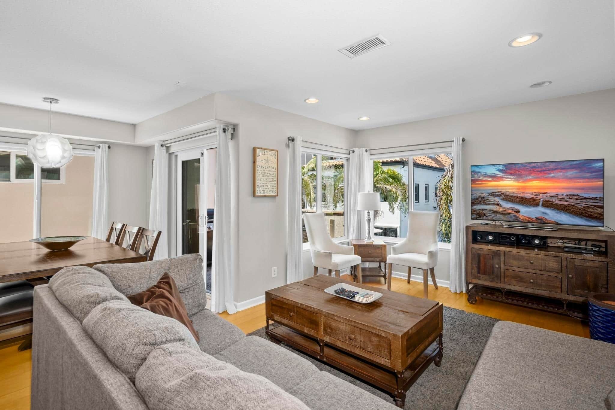 A modern living room with a sofa, coffee table, dining table, and chairs. There is a painting on the wall and a view of the outdoors through large windows.