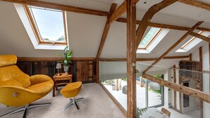 A loft-style room with exposed wooden beams, a large window, a yellow chair, a small table, and a potted plant. - 3 Bed in Ditchingham (oc-s42751) (Ditchingham)