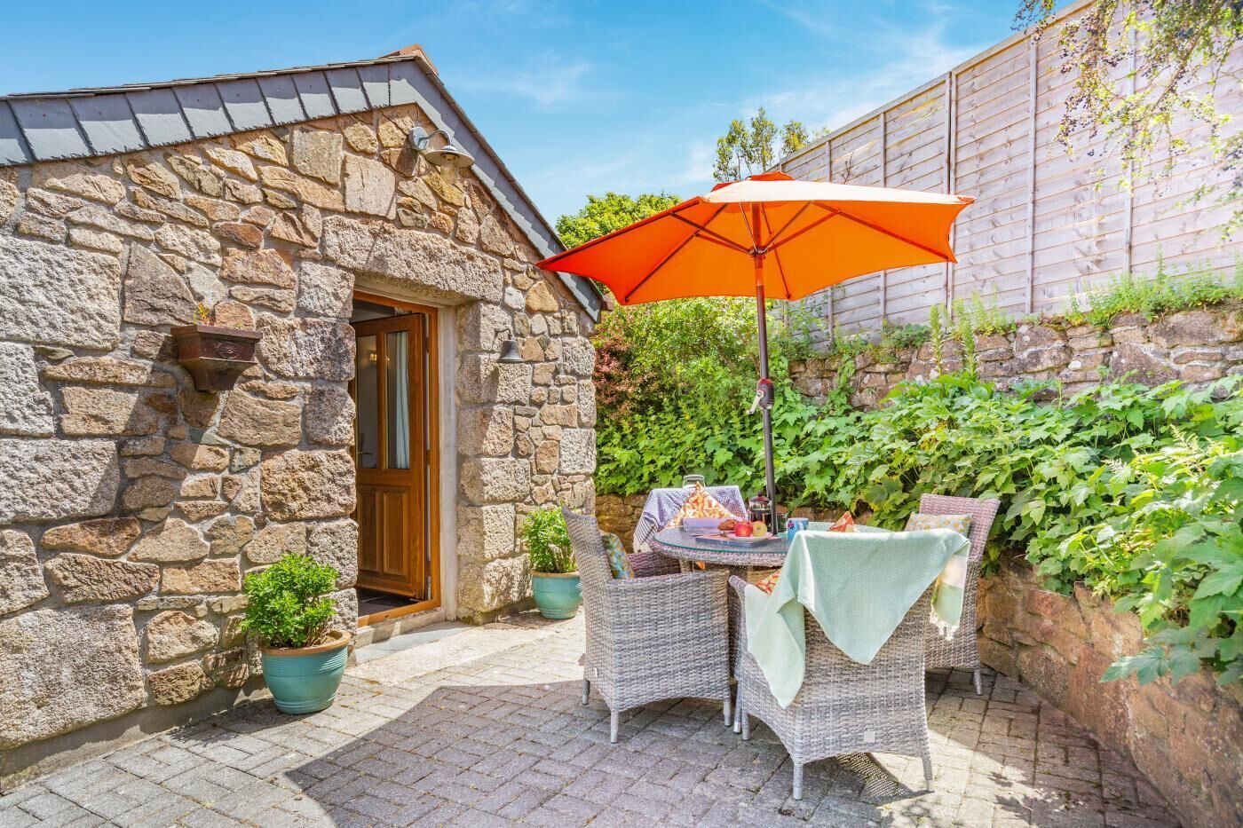 Ein Steinhaus mit einer Holztür, einer Terrasse mit Korbmöbeln und einem orangefarbenen Sonnenschirm.