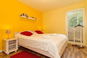 A bedroom with a bed, a nightstand, a shelf with books, a window with a view, and a red rug. - Fewo 7.0.N - Ferienwohnung in Graal-Müritz (Graal-Müritz)