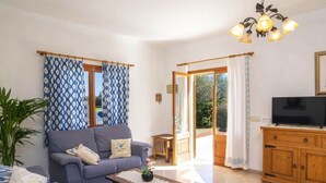A living room with a sofa, coffee table, TV, wooden cabinet, and a large window with curtains. - VILLA CAN TORRETA (Pollensa)