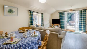 Ein gemütliches Zimmer mit Esstisch, gedeckter für ein Frühstück, einer Couch, einem Fernseher und einem Fenster mit Vorhängen.