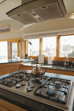 A kitchen with a gas stove, a kettle, and a window overlooking a cityscape. - Capitola Pelican House (Capitola)