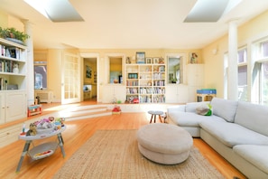 Ein geräumiges Wohnzimmer mit einer großen weißen Couch, einem kleinen Hocker, einem Bücherregal und einem Spielbereich mit Spielzeug.