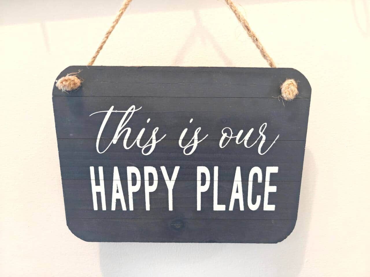 A dark wooden sign with the text 'this is our HAPPY PLACE' in white letters, hanging on a wall.