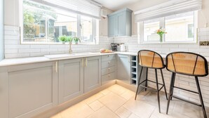 A modern kitchen with grey cabinets, a white countertop, two wicker chairs, and a window overlooking a garden. - 4 Bed in Tenby (oc-a42730) (Tenby)