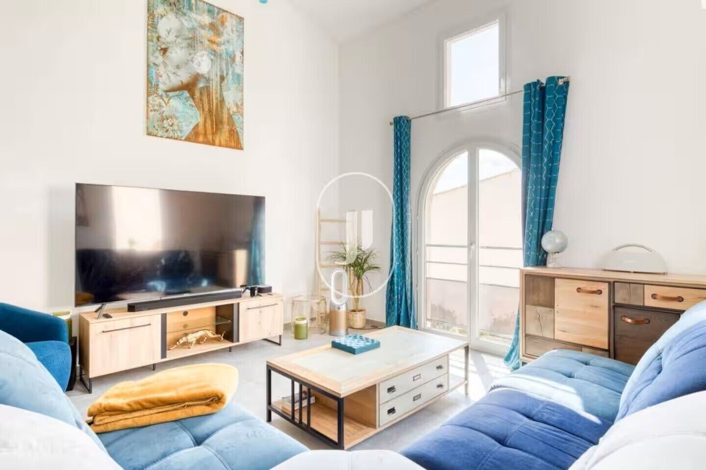 Uma sala de estar moderna com uma televisão de tela plana, sofás azuis, uma mesa de centro e uma janela grande com cortinas.